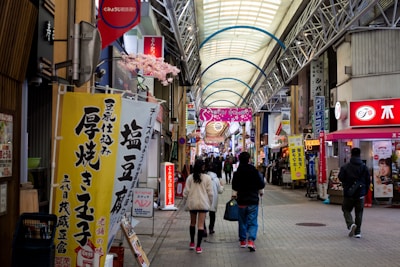 A bustling indoor shopping arcade with a variety of signs and advertisements written in Japanese. The setting features a high arched ceiling with supports and decorative elements. Shoppers are walking along the central pathway, surrounded by stores on either side offering various goods and services.