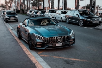 A luxury sports car is parked on the side of a street, surrounded by several other parked vehicles. The car has a sleek design with a shiny dark blue exterior and a distinctive grille. Palm trees and modern buildings are visible in the background, suggesting an urban or coastal setting.