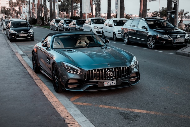 A luxury sports car is parked on the side of a street, surrounded by several other parked vehicles. The car has a sleek design with a shiny dark blue exterior and a distinctive grille. Palm trees and modern buildings are visible in the background, suggesting an urban or coastal setting.