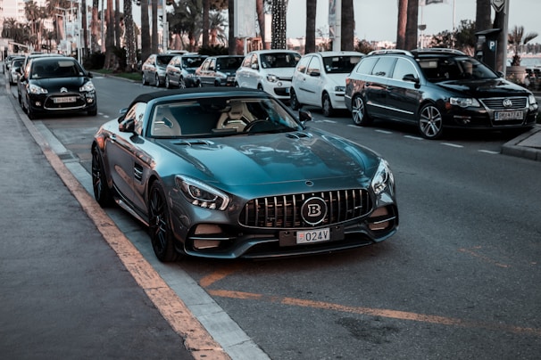A luxury sports car is parked on the side of a street, surrounded by several other parked vehicles. The car has a sleek design with a shiny dark blue exterior and a distinctive grille. Palm trees and modern buildings are visible in the background, suggesting an urban or coastal setting.