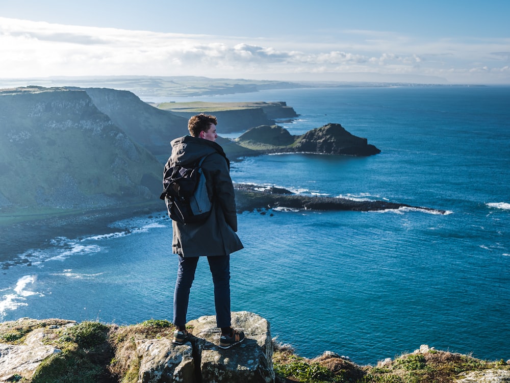 man standing on cliff looking on clear blue sea during daytime