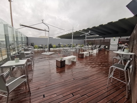An outdoor patio area with wooden flooring is set up with metal tables and chairs. There are modernistic umbrellas without any fabric, and some potted plants add greenery to the scene. The sky appears overcast, suggesting recent rain as the floor looks wet. A glass railing encloses the space on one side.