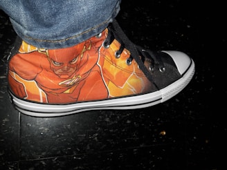 A sneaker displaying a vivid illustration of a superhero in red and orange tones with a black and white sole. The shoe features a design with eyelets for laces and stitching details. The background is a dark, textured surface.