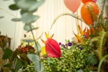 A lush display of various vibrant flowers and foliage, including a prominent red anthurium in the foreground. The background is adorned with colorful balloons in shades of orange, pink, and purple, contributing to a festive atmosphere.