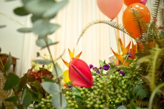 A lush display of various vibrant flowers and foliage, including a prominent red anthurium in the foreground. The background is adorned with colorful balloons in shades of orange, pink, and purple, contributing to a festive atmosphere.