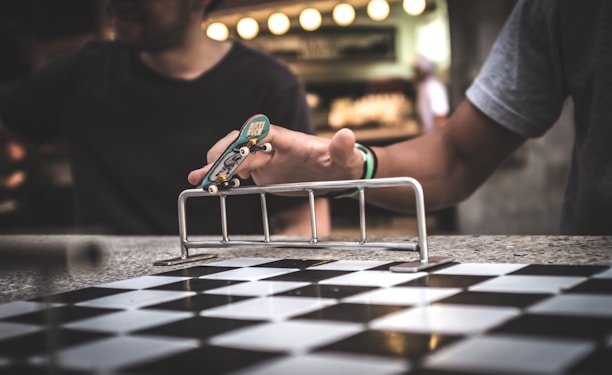 A person uses their fingers to maneuver a fingerboard skateboard over a small metal rail. The setting includes a checkered pattern surface and another person is partially visible in the background.