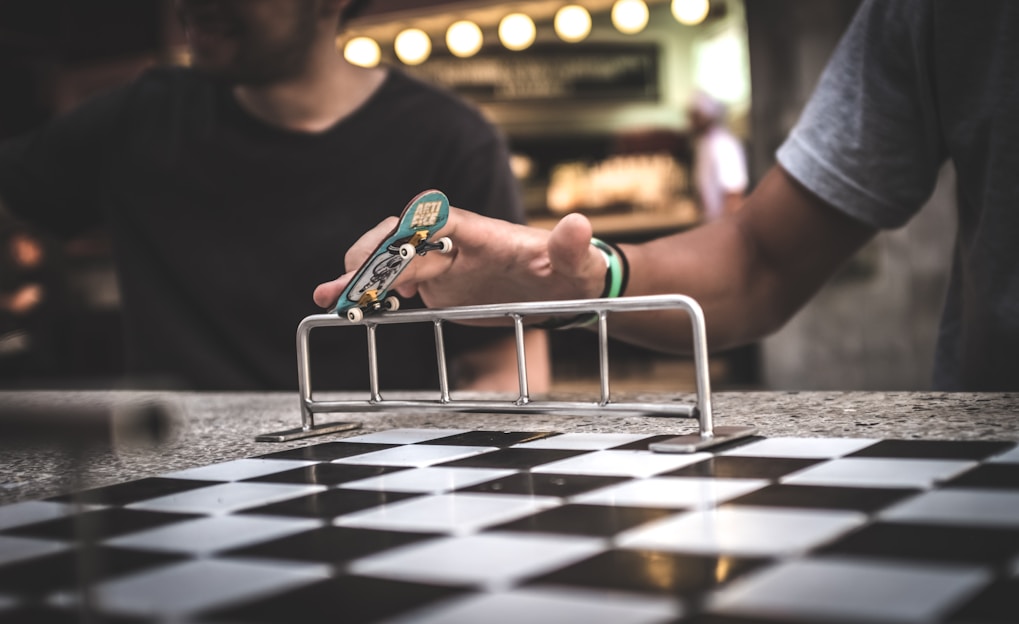 A person uses their fingers to maneuver a fingerboard skateboard over a small metal rail. The setting includes a checkered pattern surface and another person is partially visible in the background.