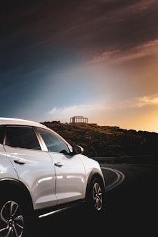 A white SUV is parked on a road with a backdrop of a lush green landscape. In the distance, an ancient temple structure stands atop a hill, surrounded by foliage. The sky above transitions from a dark, dramatic tone to a glowing warm sunset, creating a contrast between the vehicle and the natural scenery.