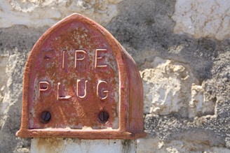 A weathered fire hydrant cap with 'FIRE PLUG' inscribed on it, featuring an aged, rusty surface. It stands against a textured stone wall, which exhibits signs of erosion and discoloration.