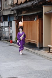 A person in traditional attire with white makeup and a purple kimono walks along a paved street. The street is lined with wooden buildings featuring bamboo blinds and a paper lantern hanging above. The setting suggests a traditional Japanese environment.