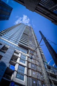 A tall, modern skyscraper is seen from a low-angle perspective, showcasing its glass facade and numerous floors. The clear blue sky with scattered clouds serves as the backdrop. The construction site is evident with visible scaffolding and building materials.