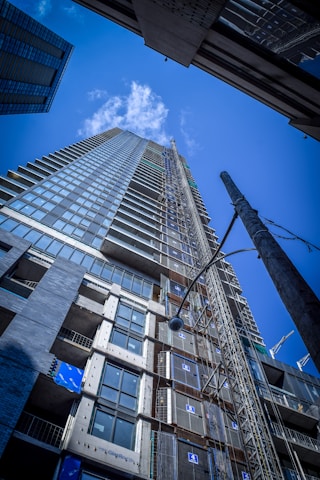 A tall, modern skyscraper is seen from a low-angle perspective, showcasing its glass facade and numerous floors. The clear blue sky with scattered clouds serves as the backdrop. The construction site is evident with visible scaffolding and building materials.