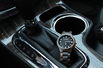 A wooden wristwatch with a dark face and simple design rests in the gear shift area of a car, highlighting the contrast between the natural wooden texture and the sleek, modern car interior.