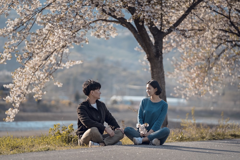 man and woman sitting on concrete road near white-leaf trees during daytime