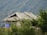 A rural thatched-roof hut is nestled among lush green foliage, with a mountainous backdrop covered in verdant vegetation. The structure is partially obscured by the surrounding greenery, giving it a secluded appearance. A blue tarpaulin is draped over one side of the building.