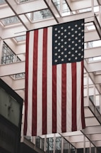 A large American flag is displayed indoors, hanging from the ceiling of a building with a modern architectural design featuring glass and metal framework.
