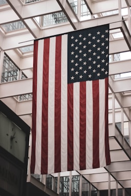 A large American flag is displayed indoors, hanging from the ceiling of a building with a modern architectural design featuring glass and metal framework.