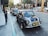 A classic Mini Cooper with a dark blue paint job and white racing stripes is parked on a city street. The car's front features yellow and white fog lights. Palm trees line the sidewalk, and pedestrians walk by, enjoying the urban scene. Modern buildings and a few other classic cars are visible in the background.