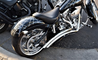 A custom motorcycle with chrome detailing and a unique spider web design on the fenders and tank is parked on a concrete street. The motorcycle features a large rear wheel and intricate engine components. Another motorcycle is partially visible in the background.