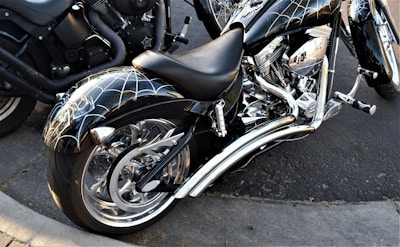 A custom motorcycle with chrome detailing and a unique spider web design on the fenders and tank is parked on a concrete street. The motorcycle features a large rear wheel and intricate engine components. Another motorcycle is partially visible in the background.