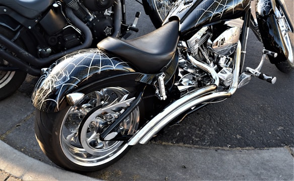 A custom motorcycle with chrome detailing and a unique spider web design on the fenders and tank is parked on a concrete street. The motorcycle features a large rear wheel and intricate engine components. Another motorcycle is partially visible in the background.