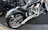 A custom motorcycle with chrome detailing and a unique spider web design on the fenders and tank is parked on a concrete street. The motorcycle features a large rear wheel and intricate engine components. Another motorcycle is partially visible in the background.