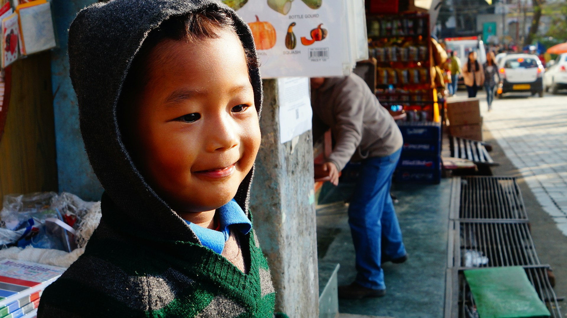 A close-up of a young child with hopeful eyes, wrapped in a worn-out sweater, standing near a busy market street in Nigeria.