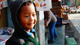 A young child wearing a hooded sweater stands near a market stall on a busy sidewalk. The child is smiling and looking content, with a warm glow from the sunlight casting soft shadows. In the background, an adult appears to be reaching into a box, while other pedestrians and parked cars are visible along the street. The market stall displays various items, including snacks and boxes.
