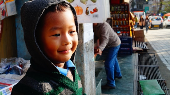 A young child wearing a hooded sweater stands near a market stall on a busy sidewalk. The child is smiling and looking content, with a warm glow from the sunlight casting soft shadows. In the background, an adult appears to be reaching into a box, while other pedestrians and parked cars are visible along the street. The market stall displays various items, including snacks and boxes.