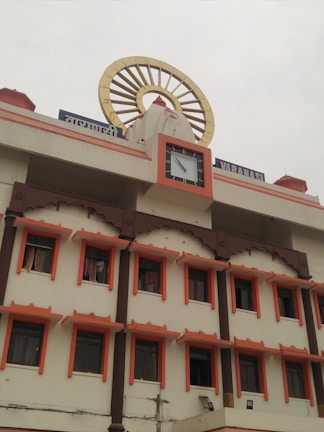 A multi-story building with a traditional architectural style featuring several windows each outlined with an orange border. On top, there's a clock and a large decorative element resembling a Dharma wheel. The name of the location is displayed in Hindi and English.