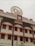 A multi-story building with a traditional architectural style featuring several windows each outlined with an orange border. On top, there's a clock and a large decorative element resembling a Dharma wheel. The name of the location is displayed in Hindi and English.