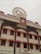 A multi-story building with a traditional architectural style featuring several windows each outlined with an orange border. On top, there's a clock and a large decorative element resembling a Dharma wheel. The name of the location is displayed in Hindi and English.