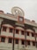 A multi-story building with a traditional architectural style featuring several windows each outlined with an orange border. On top, there's a clock and a large decorative element resembling a Dharma wheel. The name of the location is displayed in Hindi and English.