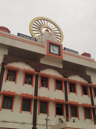 A multi-story building with a traditional architectural style featuring several windows each outlined with an orange border. On top, there's a clock and a large decorative element resembling a Dharma wheel. The name of the location is displayed in Hindi and English.
