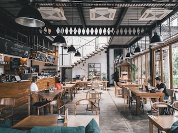 A modern caf&eacute; with a spacious, open layout. Wooden furniture, potted plants, and large windows allowing natural light to fill the room. Several people are seated, engaged in different activities such as working on laptops and conversing. Overhead, various industrial-style pendant lights hang from the ceiling. A menu board is visible above the counter where a barista prepares coffee.