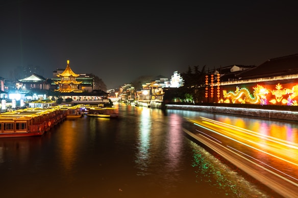 A serene night scene of a river illuminated by bright and colorful neon lights from traditional Chinese architecture. The river is lined with ornate buildings and a prominent pagoda. Light trails from boats are visible, indicating movement and activity on the water.