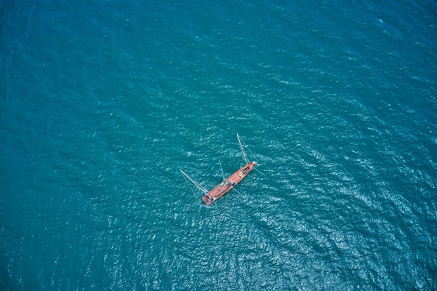 Aerial view of a solitary red sailboat sailing on a vast expanse of blue ocean water, creating a sense of isolation and adventure.