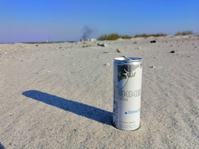 A can of energy drink is standing upright on a sandy surface. The can casts a long shadow and has a white and blue color scheme. In the background, there is a distant line of trees with some smoke rising and a clear blue sky.