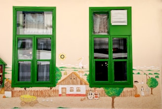 A mural painted on a wall with two green-framed windows depicting a rural scene. The artwork includes a house with a thatched roof, three figures in traditional attire sitting in front, and various farm animals such as a horse and geese. A cart filled with hay and a tree can also be seen, along with a stork standing on a rooftop next to a sun drawing.