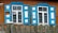 Two vintage windows with vibrant blue shutters on a wooden house wall. The windows have white and weathered frames, and a rustic wooden fence runs across the bottom part of the image.