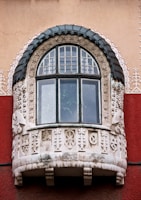 An ornately decorated window on a building, featuring intricate stone carvings and a semi-circular shape with a dark, textured frame. The carvings include geometric and abstract patterns, giving a historical or artistic appearance. The building wall is painted in a combination of beige and deep red.