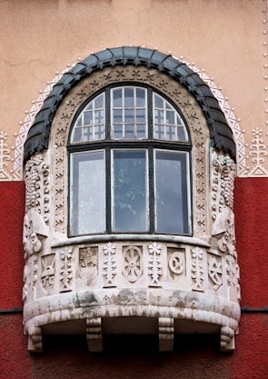 An ornately decorated window on a building, featuring intricate stone carvings and a semi-circular shape with a dark, textured frame. The carvings include geometric and abstract patterns, giving a historical or artistic appearance. The building wall is painted in a combination of beige and deep red.