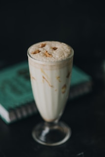 A tall glass cup filled with a creamy beverage, likely a milkshake or smoothie, with a frothy top and drizzles of caramel. It has a few almond slices on top. The glass is placed on a dark surface with a green book in the background.