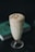 A tall glass cup filled with a creamy beverage, likely a milkshake or smoothie, with a frothy top and drizzles of caramel. It has a few almond slices on top. The glass is placed on a dark surface with a green book in the background.