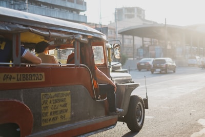 A red and silver jeepney is filled with passengers and is traveling on a road in front of a few modern buildings. The image, taken during daylight, shows a typical urban scene with vehicles and remnants of sunlight streaming in from the right side. The jeepney has signage indicating its route and there is one person seen sitting on the step at the side, almost preparing to alight.