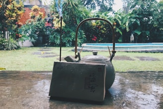 A ceramic teapot with a wooden handle sits on a wet stone surface, accompanied by a vertical incense stick and a textured block with Asian characters. In the background, a lush garden with various tropical plants surrounds a rectangular pool, creating a tranquil outdoor setting.