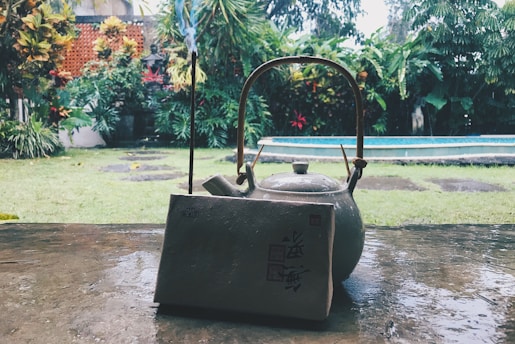 A ceramic teapot with a wooden handle sits on a wet stone surface, accompanied by a vertical incense stick and a textured block with Asian characters. In the background, a lush garden with various tropical plants surrounds a rectangular pool, creating a tranquil outdoor setting.