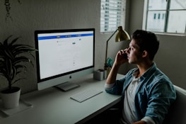 A person is sitting at a desk in front of a computer monitor displaying a financial website. The room is softly lit with natural light coming in from a nearby window. A desk lamp and a plant add to the aesthetic of the workspace. The person is holding a smartphone to their ear, appearing to be in a call.