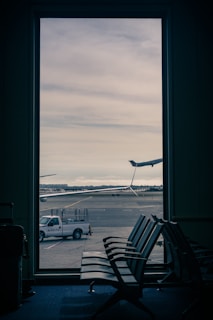 A view through a large window reveals an airport runway with a plane's tail visible in the distance. In the foreground, there are empty seats in an airport waiting area. Outside, a Delta truck is parked near the runway under a cloudy sky.