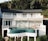 A modern white building features large windows and a curved balcony overlooking lush greenery. An infinity pool with teal tiles sits on an elevated platform. Several people are lounging on deck chairs and sitting by the poolside, enjoying the tranquil setting.